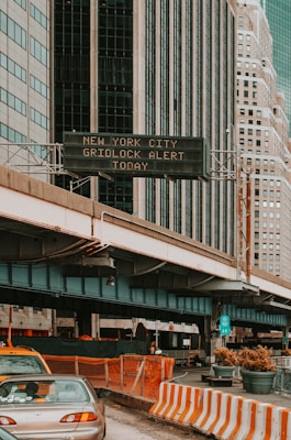 An urban scene featuring tall buildings with a digital traffic sign displaying a gridlock alert. A roadway is partially visible below with construction barriers and cars. High-rise structures dominate the background, emphasizing a city environment.