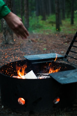 A close-up shot of a campfire pit with burning wood, surrounded by a metal ring. The flames and embers are visible, creating a warm atmosphere. A person's hand is visible above the fire, possibly warming up or cooking something. The background features a forest setting with trees and soft lighting, suggesting an outdoor adventure.