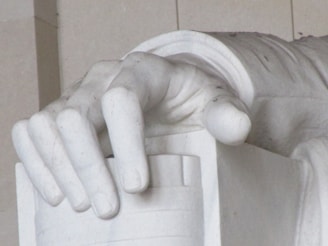 A close-up view of a large, detailed marble hand resting on an object that appears to be part of a statue. The hand is sculpted with attention to anatomical detail, displaying realistic fingers and a smooth texture typical of marble carvings.