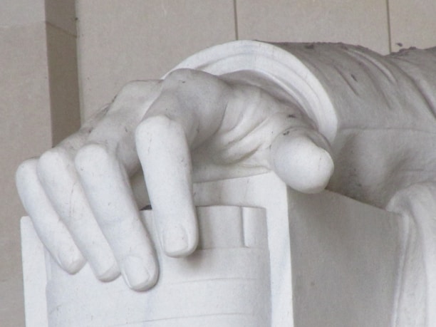 A close-up view of a large, detailed marble hand resting on an object that appears to be part of a statue. The hand is sculpted with attention to anatomical detail, displaying realistic fingers and a smooth texture typical of marble carvings.