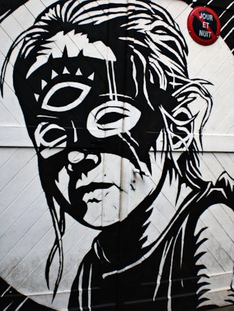 A black and white street art mural depicts a stylized face with a third eye on the forehead, surrounded by abstract patterns and lines. The face appears thoughtful and intense. A red circular sign with the words 'JOUR ET NUIT' is present above.