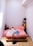 A minimalist bedroom with a wooden pallet bed frame and pink bedding. A person is lying face down on the bed, wearing light-colored shorts. There is a small side table with a potted plant and a cup on one side, and a shelving unit with a lamp and more plants on the other.