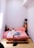 A minimalist bedroom with a wooden pallet bed frame and pink bedding. A person is lying face down on the bed, wearing light-colored shorts. There is a small side table with a potted plant and a cup on one side, and a shelving unit with a lamp and more plants on the other.