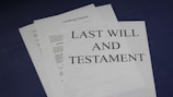 A stack of documents on a dark surface, with the top page clearly labeled 'Last Will and Testament' in bold letters. The documents appear to be legal papers, suggesting importance and formality.