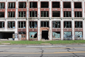A multi-story abandoned brick building with numerous broken windows and graffiti on the walls. The structure is deteriorated and appears to have been neglected for a long time. The front door is missing or severely damaged, exposing the interior. A patch of grass lies in front of the building, separated from it by a paved road.