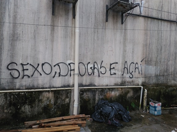 A wall features graffiti with bold, black lettering spelling out 'SEXO, DROGAS E AÇAÍ'. The surface appears weathered and discolored, with visible pipes and some hanging cables. Below the writing, there are piles of wooden planks, black plastic trash bags, and cardboard boxes on a concrete floor.