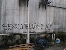A wall features graffiti with bold, black lettering spelling out 'SEXO, DROGAS E A&Ccedil;A&Iacute;'. The surface appears weathered and discolored, with visible pipes and some hanging cables. Below the writing, there are piles of wooden planks, black plastic trash bags, and cardboard boxes on a concrete floor.