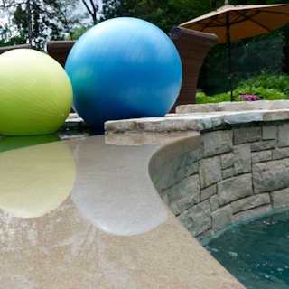 Large exercise balls in blue and green rest near the edge of a stone-tiled pool. In the background, a large patio umbrella is visible, with shrubs and a garden area adding greenery to the scene. The smooth poolside surface reflects the balls, contributing to a peaceful outdoor setting.