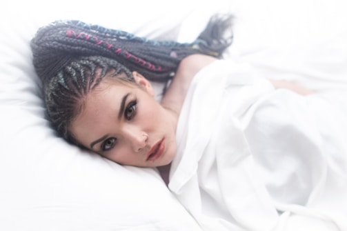 A person with intricate braided hair lies on white bedding, gazing directly at the camera. The scene is softly lit with a dreamy quality, highlighting the person's facial features and the unique style of the braids.