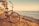 A vintage-style bicycle with orange rims is parked on a sandy beach path. Tall palm trees are lined along the path, and the ocean waves gently crash against the shore. In the background, a pier extends into the water, and the sky is painted with warm colors of late afternoon or early evening, giving a serene and peaceful atmosphere.