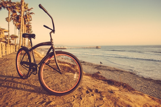 A vintage-style bicycle with orange rims is parked on a sandy beach path. Tall palm trees are lined along the path, and the ocean waves gently crash against the shore. In the background, a pier extends into the water, and the sky is painted with warm colors of late afternoon or early evening, giving a serene and peaceful atmosphere.