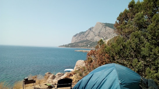 A serene coastal landscape with a distant mountain, a calm blue sea, and a campsite setup including a blue tent, a chair, and a small table. Lush greenery partially obscures the view to the right.