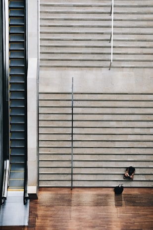 A broad staircase is juxtaposed next to an escalator, with a person sitting alone on the steps alongside a bag. The stark contrast between the static stairs and the moving escalator highlights themes of choice and solitude.