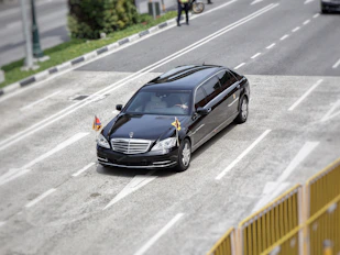A black luxury car, possibly a limousine, travels on a multi-lane road with small flags attached to its front. The setting includes some greenery and a person on a bicycle in the background.