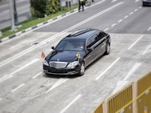 A black luxury car, possibly a limousine, travels on a multi-lane road with small flags attached to its front. The setting includes some greenery and a person on a bicycle in the background.