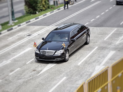 A black luxury car, possibly a limousine, travels on a multi-lane road with small flags attached to its front. The setting includes some greenery and a person on a bicycle in the background.
