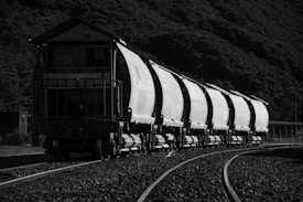 A long train composed of cargo wagons travels on tracks curving through a mountainous area. The image is in black and white, highlighting the contrast between the industrial nature of the train and the natural landscape surrounding it.