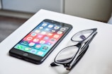 A smartphone with a colorful display of app icons is placed on a white surface next to a pair of black-framed eyeglasses.