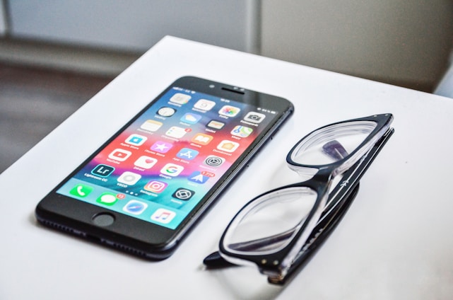 A smartphone with a colorful display of app icons is placed on a white surface next to a pair of black-framed eyeglasses.