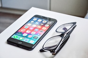 A smartphone with a colorful display of app icons is placed on a white surface next to a pair of black-framed eyeglasses.