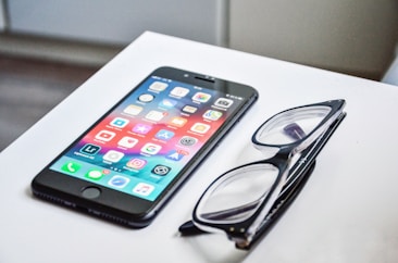 A smartphone with a colorful display of app icons is placed on a white surface next to a pair of black-framed eyeglasses.