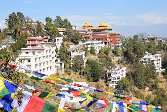 A hillside scene featuring several colorful buildings with traditional architectural elements, surrounded by lush greenery. Multicolored prayer flags are prominently displayed in the foreground, while snow-capped mountains are visible in the background.