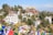 A hillside scene featuring several colorful buildings with traditional architectural elements, surrounded by lush greenery. Multicolored prayer flags are prominently displayed in the foreground, while snow-capped mountains are visible in the background.
