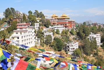 A hillside scene featuring several colorful buildings with traditional architectural elements, surrounded by lush greenery. Multicolored prayer flags are prominently displayed in the foreground, while snow-capped mountains are visible in the background.