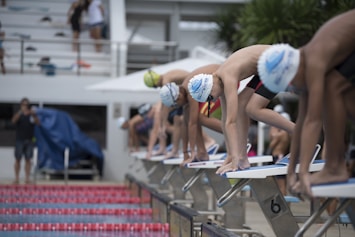 Young swimmers in swim caps and goggles are poised on starting blocks, preparing for a race. The setting is an outdoor swimming pool with a crowd in the background, creating an atmosphere of anticipation and focus.