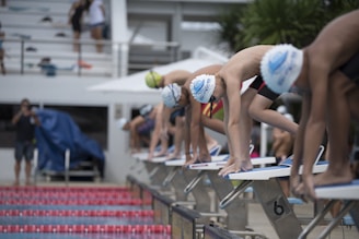 Young swimmers in swim caps and goggles are poised on starting blocks, preparing for a race. The setting is an outdoor swimming pool with a crowd in the background, creating an atmosphere of anticipation and focus.
