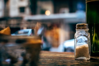 A clear glass salt shaker filled with salt is placed on a wooden surface. The background is blurred, showing out-of-focus lights and shapes, suggesting an indoor or outdoor dining setting.