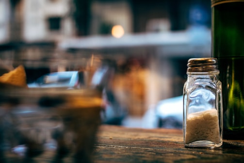 A clear glass salt shaker filled with salt is placed on a wooden surface. The background is blurred, showing out-of-focus lights and shapes, suggesting an indoor or outdoor dining setting.
