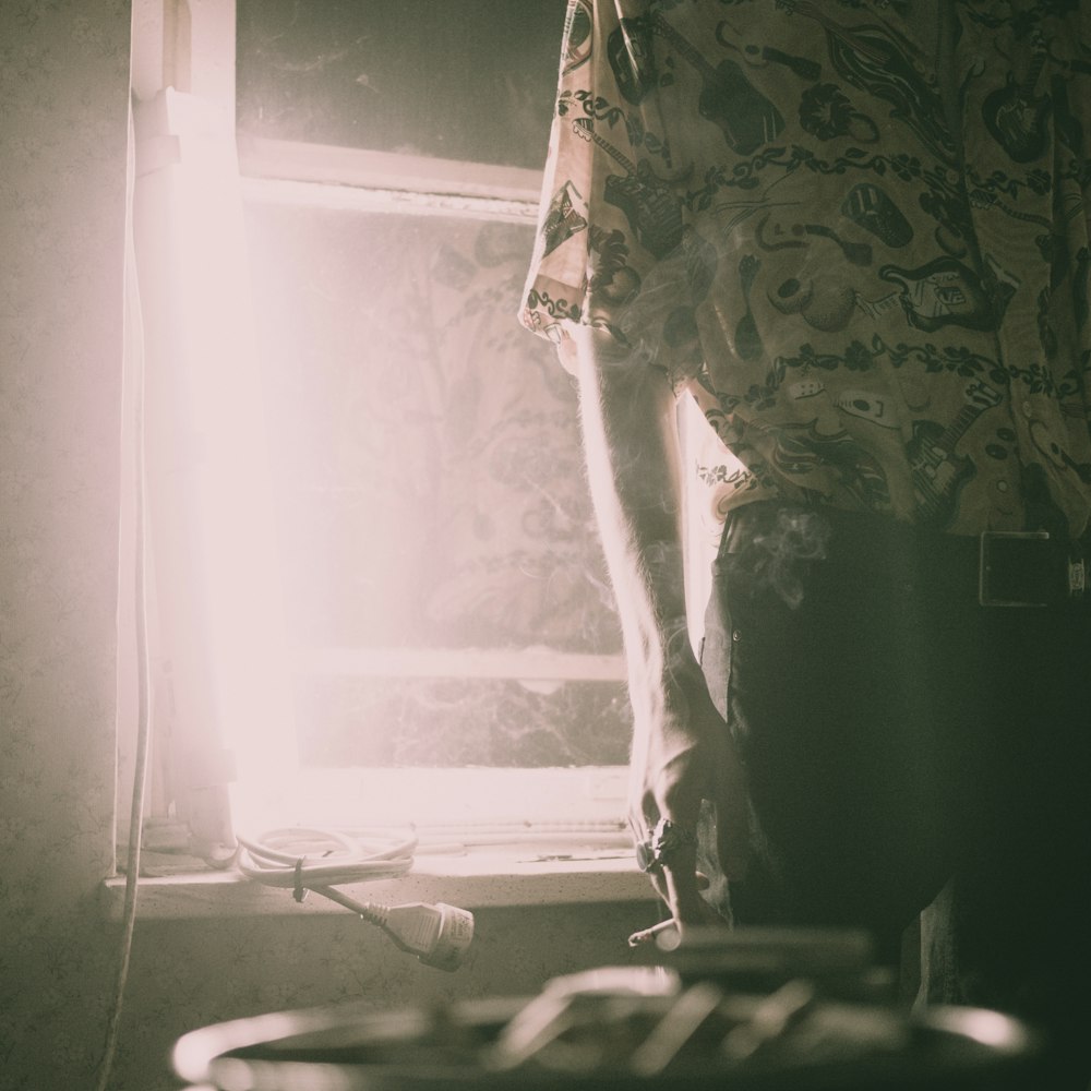 person holding cigarette standimg beside window inside the house