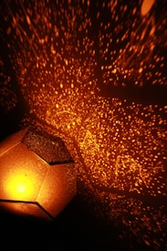 A geometric lamp projects constellations and stars onto a wall in a dark room. The lamp emits a warm orange light, creating a cozy and magical atmosphere. The star patterns appear intricate and cover a large area, adding to the visual complexity.