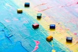 A canvas covered with overlapping vibrant colors such as blue, yellow, pink, and green, with a textured surface. Several small, square oil pastels or tiles are scattered on top of the painted canvas, adding a geometric element to the abstract background.
