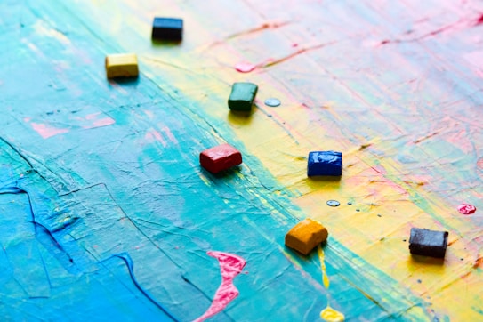 A canvas covered with overlapping vibrant colors such as blue, yellow, pink, and green, with a textured surface. Several small, square oil pastels or tiles are scattered on top of the painted canvas, adding a geometric element to the abstract background.