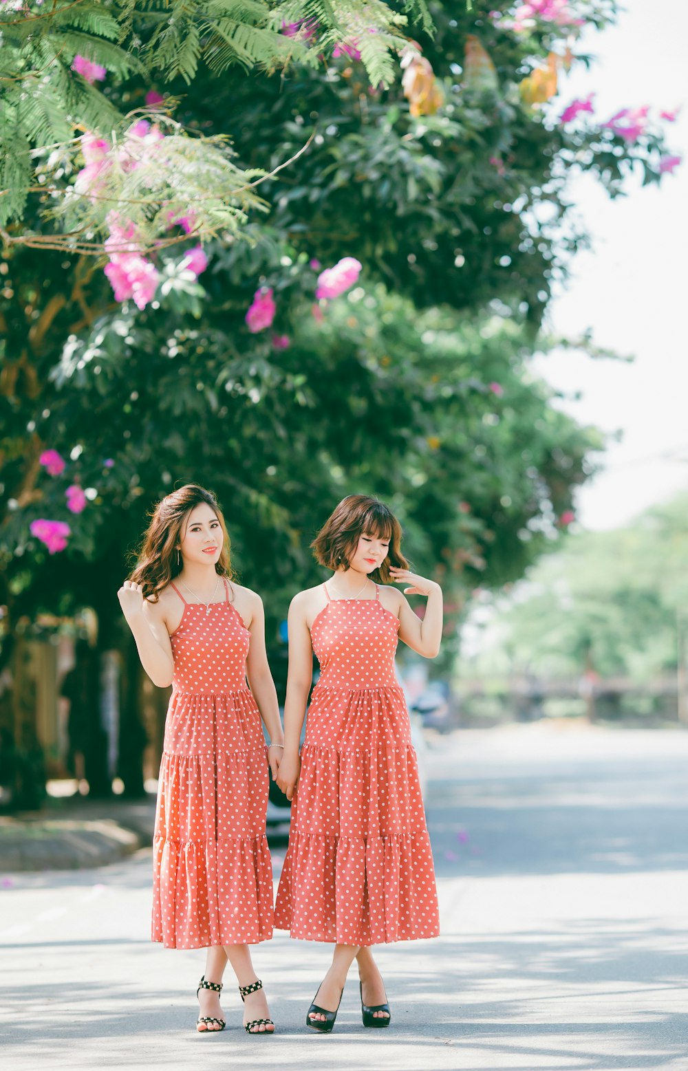 빨간색과 흰색의 물방울 무늬 드레스를 입은 두 여성이 낮에 분홍색 꽃잎이 달린 꽃 근처 도로에 서 있습니다