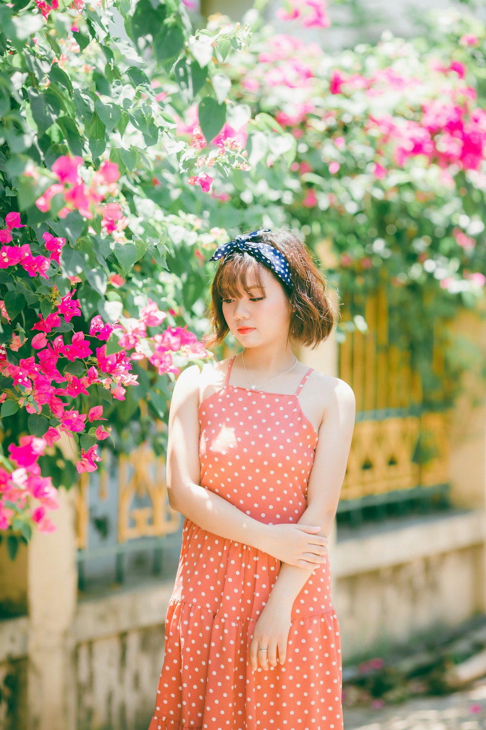 Mujer en vestido de tirantes de espagueti de lunares naranjas de pie cerca de plantas rosas y verdes