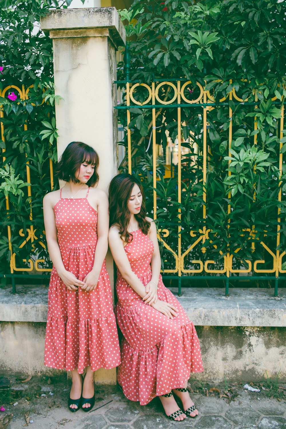 two women wearing red-and-white polka-dot spaghetti strap dress leaning on gray concrete pillar