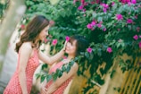 Two women in matching polka dot dresses are smiling at each other surrounded by vibrant pink flowers and lush green leaves. One woman gently touches the other's face, creating a warm and affectionate interaction.