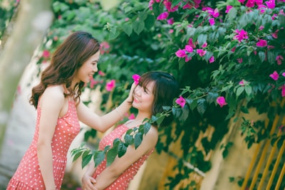 Two women in matching polka dot dresses are smiling at each other surrounded by vibrant pink flowers and lush green leaves. One woman gently touches the other's face, creating a warm and affectionate interaction.