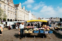 A vibrant outdoor market scene set against a backdrop of historic European architecture. The market stalls are covered with yellow and white striped canopies and display an array of colorful flowers in baskets. People are walking and browsing the stalls, and a few historic buildings with ornate fa&ccedil;ades and gabled roofs line the cobblestone street. The sky is clear with a few scattered clouds, suggesting a bright, sunny day.