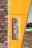 A silver intercom system is mounted on a bright yellow panel that is part of a building's exterior wall. The structure consists of light brown bricks on the left side. A white car is parked on a paved surface in the background.