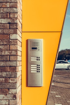 A silver intercom system is mounted on a bright yellow panel that is part of a building's exterior wall. The structure consists of light brown bricks on the left side. A white car is parked on a paved surface in the background.