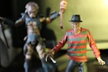 Two detailed action figures are in focus. The foreground figure has a burnt face, wearing a fedora and a red and green striped sweater, characterized by sharp claws on one hand. The background figure appears monstrous, with textured skin and holding a weapon in each hand.