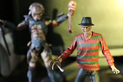 Two detailed action figures are in focus. The foreground figure has a burnt face, wearing a fedora and a red and green striped sweater, characterized by sharp claws on one hand. The background figure appears monstrous, with textured skin and holding a weapon in each hand.