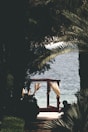 A shaded path surrounded by palm trees leads to a pergola with flowing white curtains, overlooking a tranquil sea. The scene conveys a sense of secluded relaxation, with the sunlight softly illuminating the curtains against the backdrop of the ocean.