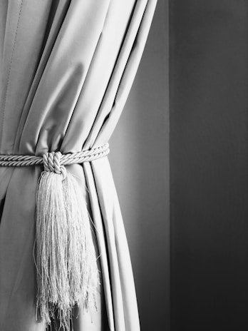 A close-up view of a curtain tied back with a braided tassel cord. The curtain fabric appears smooth and heavy, while the tassel adds a touch of elegance with its intricate braid and flowing strands. The image is in black and white, emphasizing the texture and contrast between the fabric and the background.