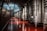 A series of large stainless steel fermentation tanks line the right side of a spacious, dimly lit industrial room. The floor has a polished, reddish-brown surface that reflects the light coming from above. The walls are curved and metallic, suggesting a tunnel-like environment.