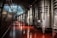 A series of large stainless steel fermentation tanks line the right side of a spacious, dimly lit industrial room. The floor has a polished, reddish-brown surface that reflects the light coming from above. The walls are curved and metallic, suggesting a tunnel-like environment.
